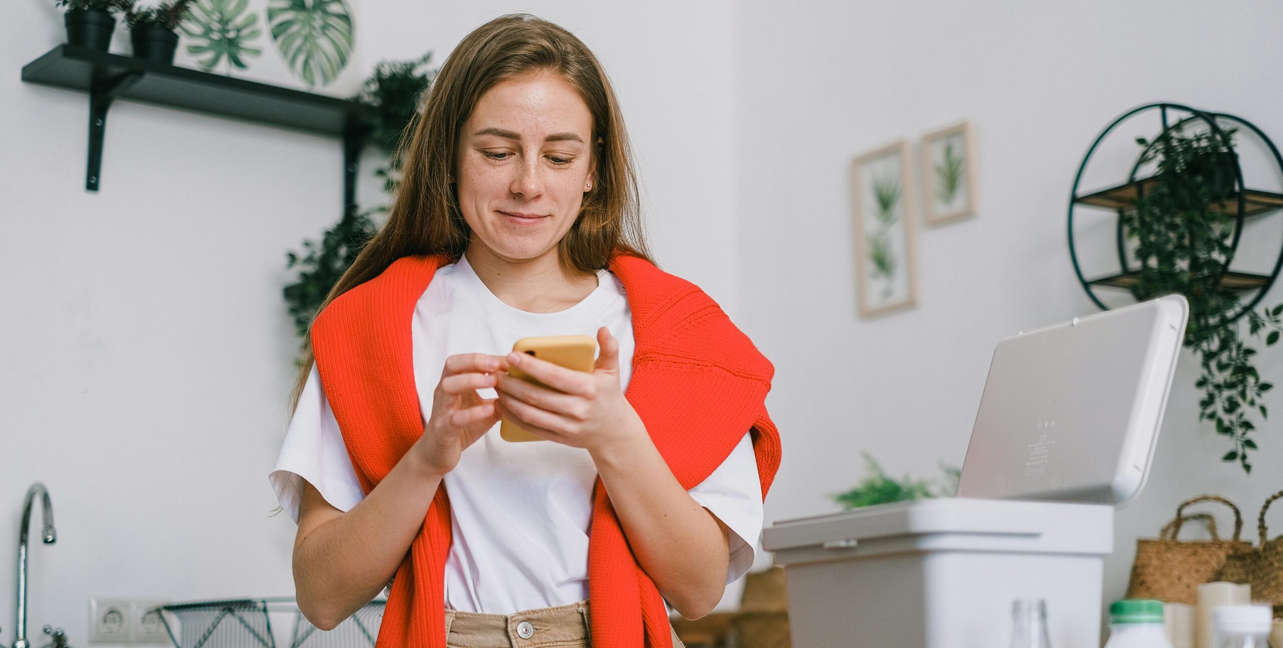Cheerful female browsing mobile phone and standing at table with sorted paper and plastic waste